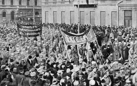 Multitud con pancartas y uniformes militares marchando en una calle, con edificios al fondo.