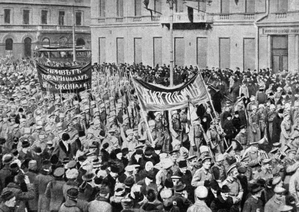 Multitud con pancartas y uniformes militares marchando en una calle, con edificios al fondo.