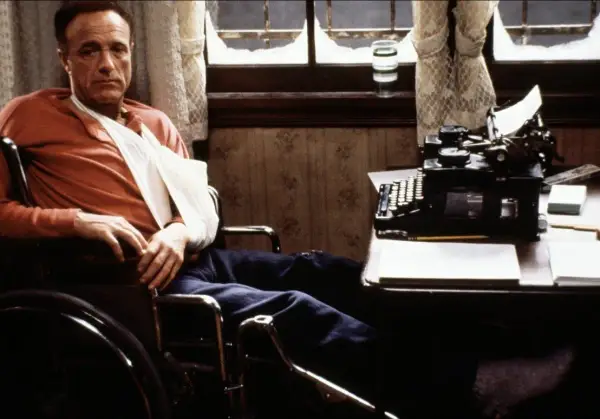 Un escritor en silla de ruedas, con un brazo en cabestrillo, mira fijamente una máquina de escribir. Escenas como esta son comunes en películas sobre escritores.